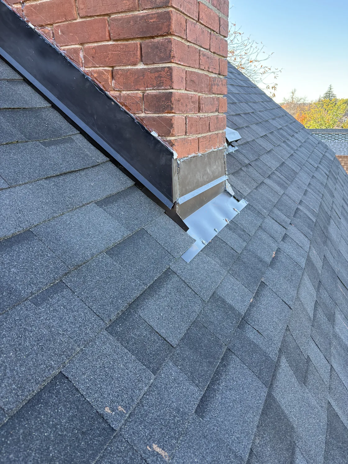 Close view of a brick chimney, metal flashing and surrounding gray roof shingles.