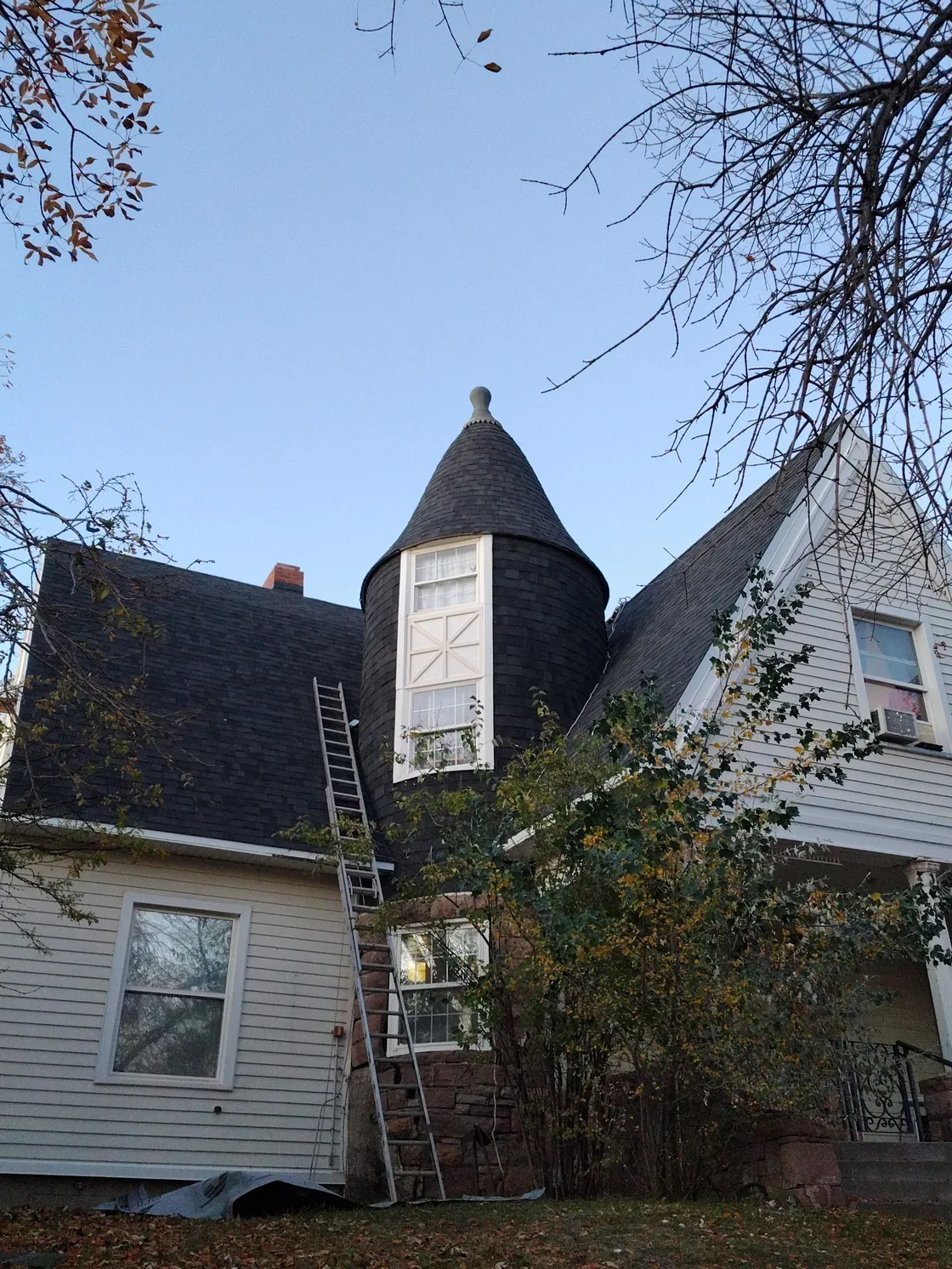 Dark shingle roofing on a conical turret and adjoining house roof, with a ladder at the roof edge.